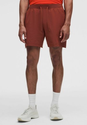 Mann trägt rostfarbene Shorts mit Kordelzug, weiße Socken und weiße Sportschuhe und steht vor einem schlichten hellen Hintergrund.