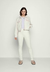 Jaqueta de denim branca com costuras rosas, combinada com uma blusa lilás clara e jeans skinny brancos. Usada com sandálias em tom de pele com detalhes em prata.