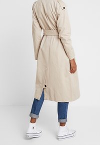 Trench coat beige con cintura, dotato di apertura posteriore. Abbinato a jeans blu con risvolto e sneakers bianche alte. Texture liscia.