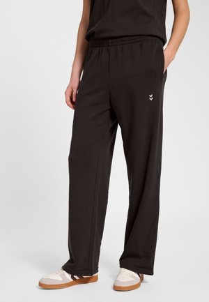 Osoba ubrana w czarne luźne spodnie z elastycznym pasem i białe trampki z brązowymi podeszwami, stojąca z jedną ręką w kieszeni.