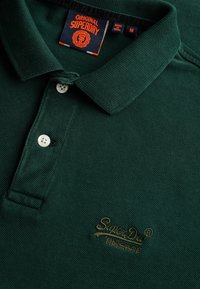 Polo shirt verde scuro realizzato in tessuto testurizzato, con colletto, due bottoni bianchi e un logo ricamato dorato.