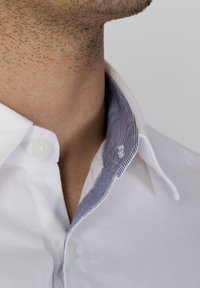 Camicia bianca con colletto bottonato, caratterizzata da una fodera interna a righe blu e bianche. Primo piano sulla zona del colletto, che rivela una texture liscia e cuciture accurate.