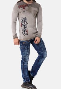 Camiseta henley gris de manga larga con bolsillo en el pecho y texto gráfico en rojo y negro. Pantalones cargo azules con múltiples bolsillos y paneles texturizados. Zapatos negros.