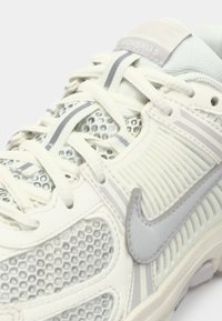 Sapatilha atlética branca com parte superior em malha, detalhes cinzentos e detalhes texturizados. Apresenta um sistema de atacadores, colarinho acolchoado e sola de borracha.