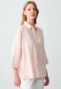 Camicia button-up rosa chiaro a maniche corte con colletto. Tessuto testurizzato, vestibilità rilassata, con bottoni frontali e polsini arrotolati.