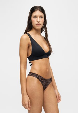 Kvinde med langt mørkt hår iført en sort bikini top og brune bikini underdele med leopardprint, stående mod en ensfarvet lys baggrund.