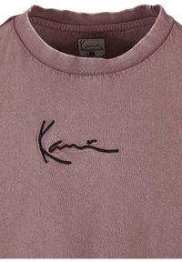 Lühikeste varrukatega lilla puuvillane T-särk ümmarguse kaelusega ja musta tikandiga logo rinnal. Tekstuur näeb välja pehme ja veidi tuhm.