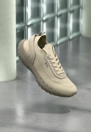 Beige gestrickter Sneaker mit dicker, strukturierter Sohle und Schnürsenkeln, der über einem grauen Boden schwebt, im Hintergrund große Fensterblöcke.