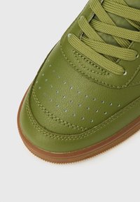 Približni prikaz prsta zelene kožne tenisice s perforiranim rupicama, zelenim vezicama u istoj boji i smeđim gumenim đonom.