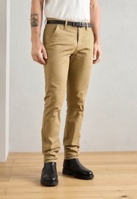 Beige chinos med smal passform, med side lommer og belte, kombinert med svarte ankelstøvler på et tregulv.
