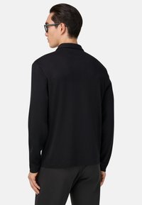 Melnas garām piedurknēm polo krekls ar plakanu apkakli, kam ir gluda tekstūra un brīva piegriezuma forma. Aizmugurējā kakla daļā ir diskrēta logotipa detaļa.