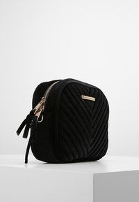 Černá sametová kosmetická taška s prošíváním, se zlatým zipem a táhly, stojící na bílé ploše na pozadí s jemným světlým tónem.