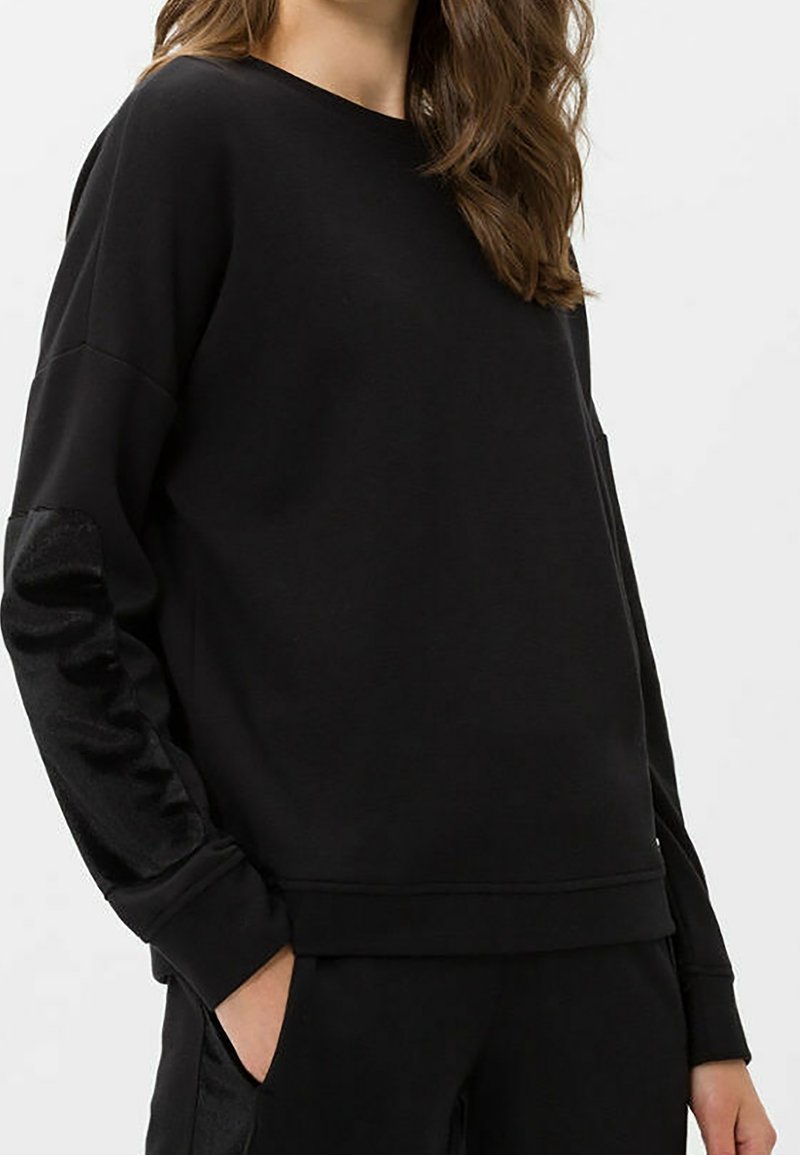 Femme portant un sweat-shirt noir à manches longues avec des coudières texturées et un pantalon noir assorti, les mains dans les poches.