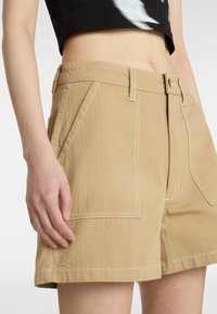 Shorts color caqui con bolsillos frontales, pasadores para cinturón y costuras blancas visibles sobre una tela texturizada, mostrados sobre una mano que descansa cerca de la cinturilla.
