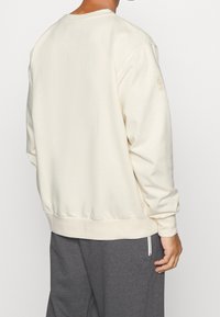 Cremefarvet sweatshirt lavet af blødt stof, med ribkant ved kanten og manchetterne, samt en logoaccent på venstre ærme.