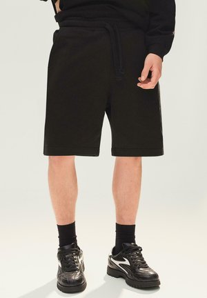 Férfi fekete térdig érő rövidnadrágban, fekete zokniban és fekete sportcipőben, egy egyszerű világos háttér előtt állva.