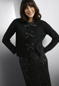 Cardigan en tricot noir avec trois nœuds en satin noir comme fermetures. Associé à une jupe noire présentant un motif géométrique en sequins.