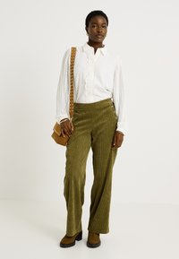 Blusa blanca texturizada con detalle de encaje, combinada con pantalones de pana verde de pierna ancha con botones laterales. Bolso cruzado marrón y botas de tobillo.
