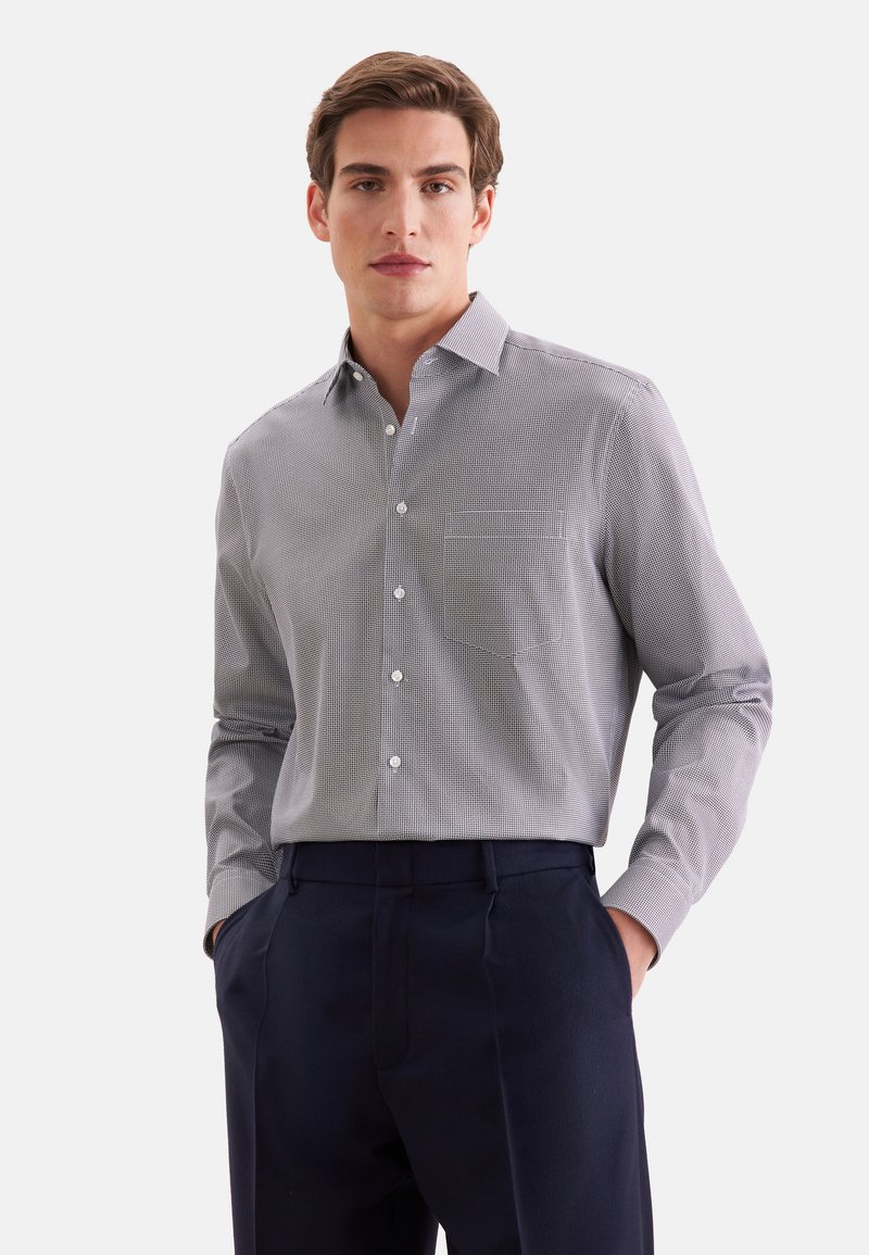 Jeune homme portant une chemise grise à motifs boutonnée, rentrée dans un pantalon bleu marine foncé, debout les mains dans les poches sur un fond uni.