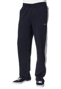Černé sportovní tepláky Adidas s bílými třemi pruhy po stranách, nošené s černobílými teniskami na stojící osobě.