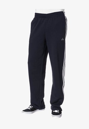 Zwarte Adidas trainingsbroek met witte drie-strepen aan de zijkant, gedragen met zwart-witte sneakers door een persoon die staat.
