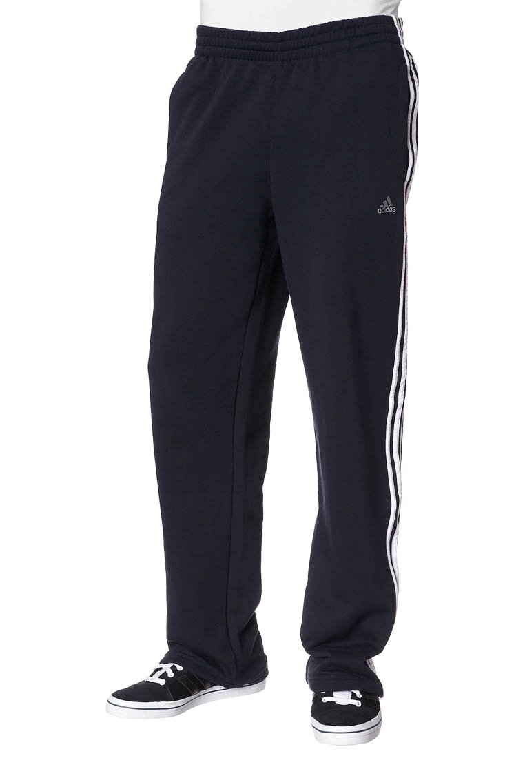 Černé sportovní tepláky Adidas s bílými třemi pruhy po stranách, nošené s černobílými teniskami na stojící osobě.
