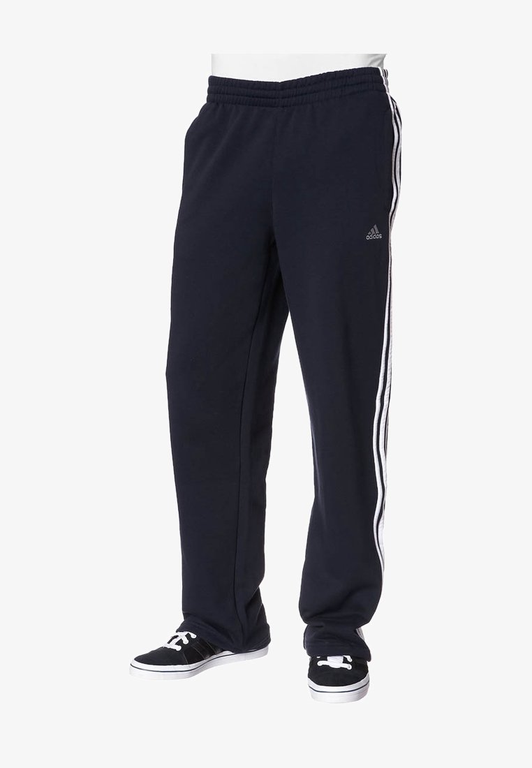 Černé sportovní tepláky Adidas s bílými třemi pruhy po stranách, nošené s černobílými teniskami na stojící osobě.