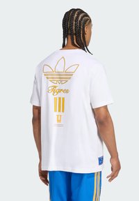 Witte katoenen T-shirt met een grote gouden Adidas-logo en "Tigres" gedrukt op de achterkant, met een ontspannen pasvorm en korte mouwen.