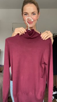 Donna che tiene un maglione a collo alto bordeaux su una gruccia bianca in una stanza con una porta chiusa e un appendiabiti visibile.