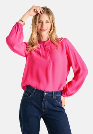 Frau mit blondem Haar, die eine knallpinkfarbene Langarmbluse und dunkelblaue Jeans trägt, lächelt und berührt mit einer Hand ihr Haar.