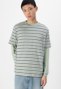 Camiseta de manga corta a rayas en verde, blanco y azul marino; cuenta con un bolsillo en el pecho. Llevada sobre una camisa de manga larga verde. Algodón ligero.