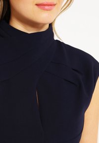 Granatowa sukienka z wysokim kołnierzem, z drapowanym materiałem, plisami i detalem w kształcie dziurki. Wykonana z usztywnionego materiału o gładkiej fakturze.