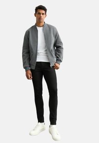 Grå bomberjacka med strukturerad yta, bärs över en vit t-shirt, tillsammans med slim fit svarta byxor och vita sneakers.