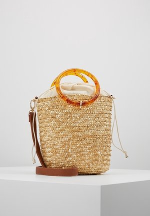 Sac seau en paille tressée avec fermeture à cordon en tissu beige, poignées circulaires en ambre et bandoulière en cuir marron sur surface blanche.