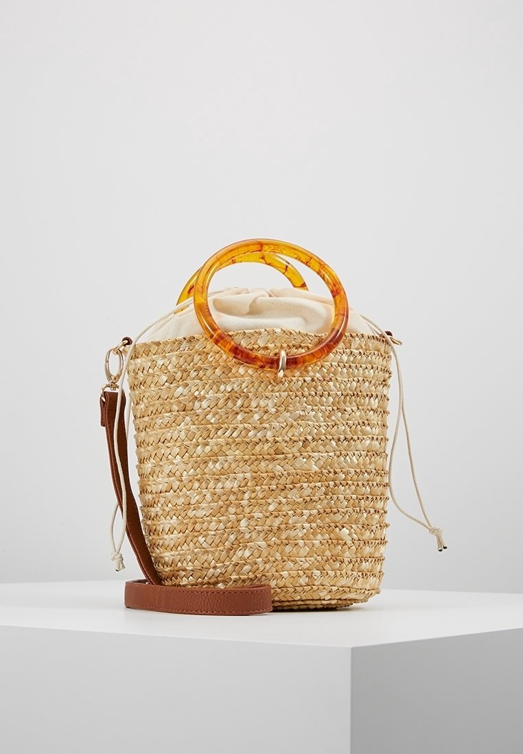 Sac seau en paille tressée avec fermeture à cordon en tissu beige, poignées circulaires en ambre et bandoulière en cuir marron sur surface blanche.