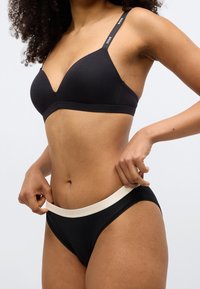 Μαύρο ασύρματο σουτιέν με V-λαιμόκοψη και λεπτές τιράντες. Συνοδευτικό μαύρο εσώρουχο με κρεμ ζώνη μέσης με λογότυπο.