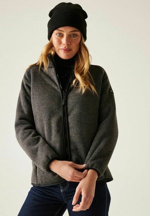 Femme portant un bonnet noir, une veste zippée gris foncé, un col roulé noir et un jean bleu foncé, posant avec les mains près de l'ourlet de la veste.