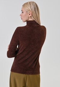 Maglione marrone peloso con collo alto e maniche lunghe, caratterizzato da una texture morbida e una vestibilità comoda, abbinato a pantaloni verde oliva.