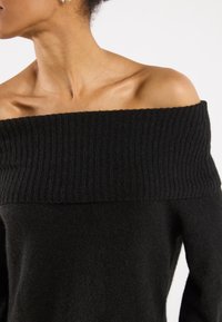 Černý svetr bez ramenních švů s žebrovaným výstřihem a měkkou texturou, odhalující horní část hrudníku a ramena.