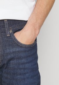 Tmavě modré džíny s úzkým střihem, se žlutou nití, kovovým knoflíkem a přední kapsou, do které je ruka vložena.