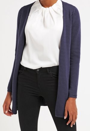 Nainen, joka pukeutunut valkoiseen pliseerattuun paitapuseroon, tummansiniseen ribbineuleiseen avoimeen neuletakkiin ja mustiin istuviin housuihin, seisoo valkoista taustaa vasten.