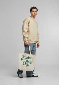 Geantă din pânză tip tote cu textul "Clubul de alergare de duminică" imprimat în verde, purtată de un model în pulover bej și blugi albaștri. Pantofi sport gri la picioare.