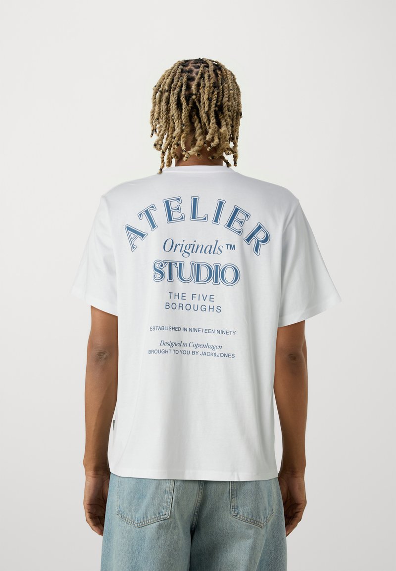 Λευκό βαμβακερό t-shirt με μπλε γραφικά κείμενα στην πλάτη, που διαθέτει "ATELIER Originals STUDIO" και επιπλέον λεπτομέρειες branding από κάτω.