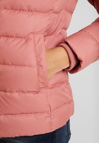 Main dans la poche d'une veste matelassée rose avec un poignet côtelé, portée sur un jean en denim bleu sur un fond uni.