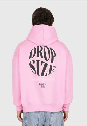 Person in einem hellrosa Hoodie mit der Aufschrift "DROP SIZE" in großen schwarzen Buchstaben und kleinerem Text darunter, kombiniert mit hellblauen Jeans.