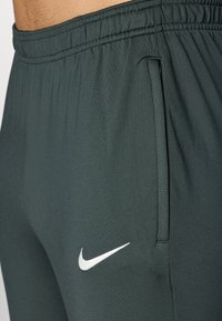 Mørkegrønne træningsbukser med en elastisk talje, sidelommer og et hvidt Nike-logo nederst til højre. Glat, letvægtsstof.