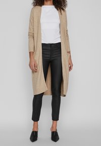 Cardigan beige en tricot avec poignets côtelés, porté sur une chemise blanche, associé à un pantalon en cuir noir et des chaussures noires à bout pointu.