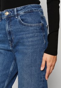 Detailní záběr na osobu v modrých denimových džínách s vysokým pasem, se zlatým knoflíkem a černým topem s dlouhým rukávem, ruka opřená o stehno.