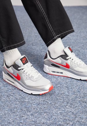 Person trägt weiße Nike Air Max Sneakers mit roten Swoosh-Logos, weißen Socken und schwarzen Hosen und steht auf einem blauen Teppichboden.