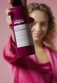L'Oréal Curl Expression sprayfles, cilindervorm, glanzend donkerroze, gelabeld met witte tekst, 120 ml. Achtergrond is zacht roze.